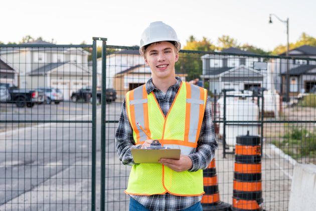 Young man in a hard hat and orange vest with a clip board smiling at the camera.
